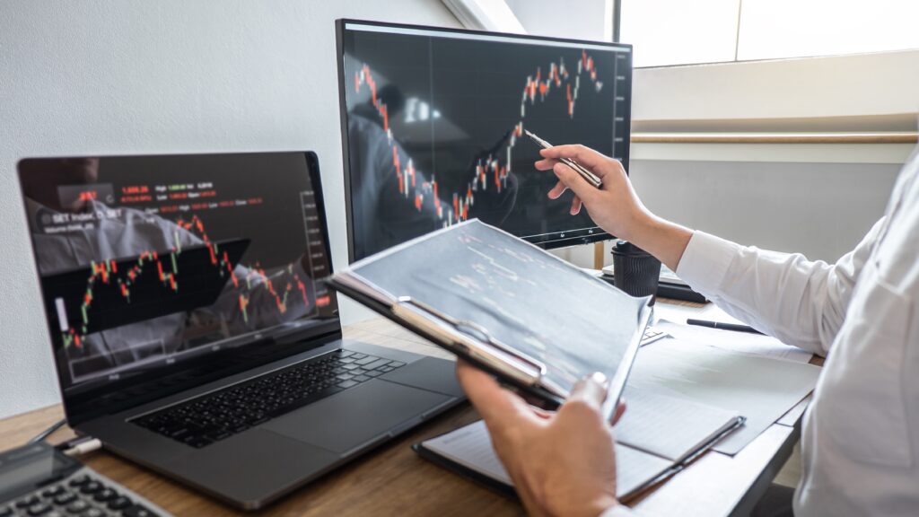 Person analyzing stock market charts on two screens with a pen in hand, holding a clipboard.